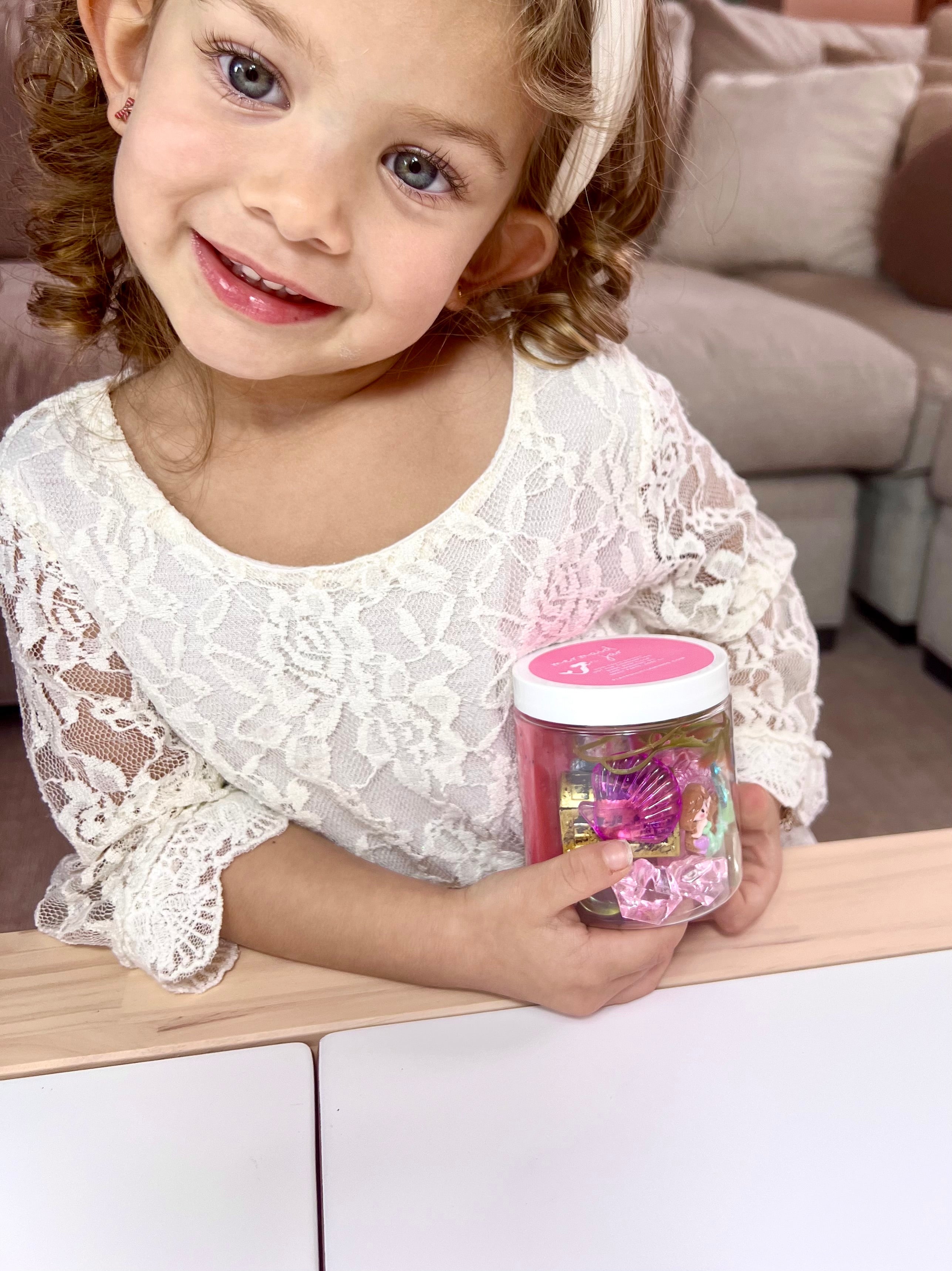 Young girl smiling and holding a jar of colorful sensory playdough pieces from the Enchanting Mermaid Playdough Sensory Kit while sitting at a table in a cozy living room.