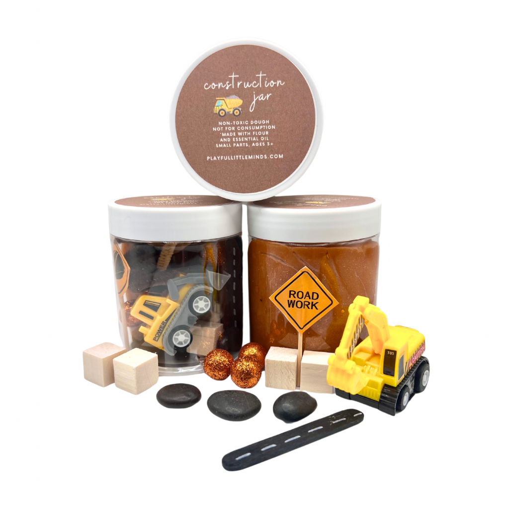Construction Site Playdough Sensory Kit | Natural Dough with Trucks, Rocks & Gems featuring two jars of brown playdough, toy construction trucks, a road work sign, wooden blocks, black stones, and decorative gems for sensory play.