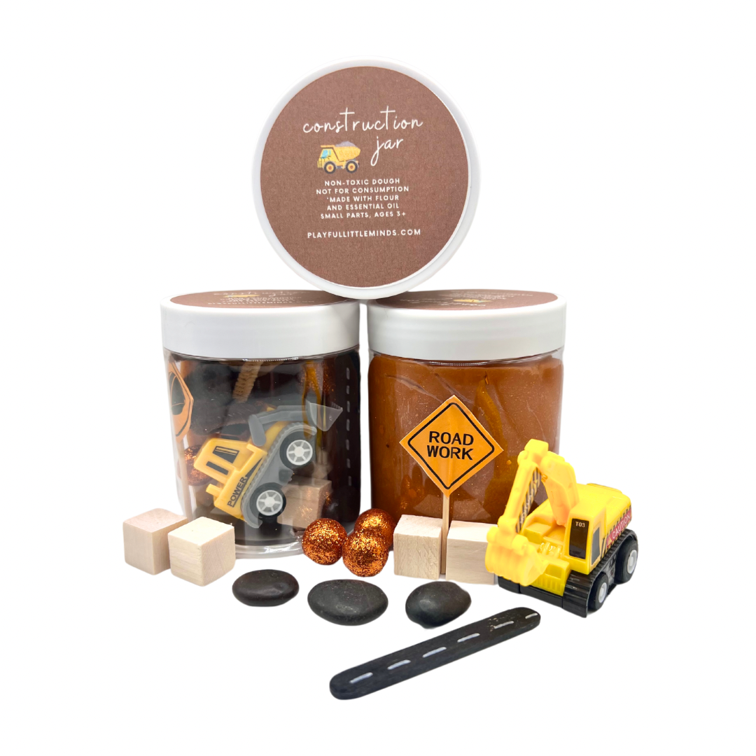 Construction Site Playdough Sensory Kit | Natural Dough with Trucks, Rocks & Gems featuring two jars of brown playdough, toy construction trucks, a road work sign, wooden blocks, black stones, and decorative gems for sensory play.