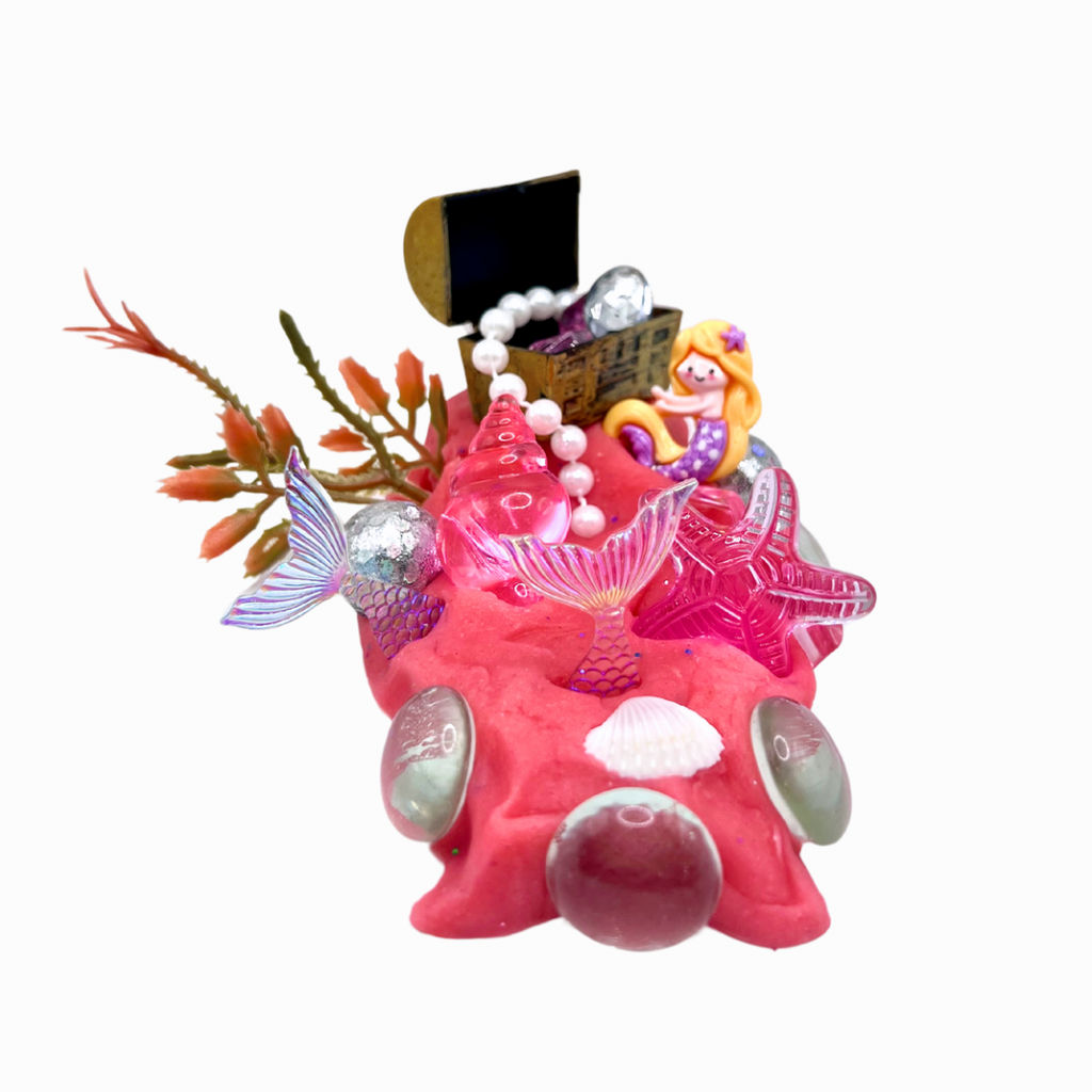 Bright pink playdough decorated with iridescent shells, a treasure chest, pearl beads, seaweed, and a small mermaid figure from the Enchanting Mermaid Playdough Sensory Kit.