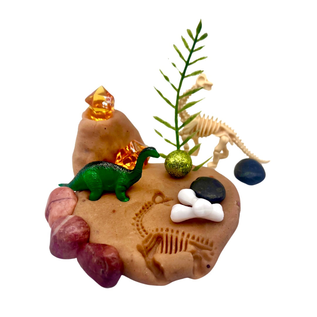 Dinosaur Playdough Sensory Kit | Natural Dough with Dino Fossils & Surprises featuring green and beige dinosaur toys, fossil imprints, decorative stones, and small plant elements on brown dough.
