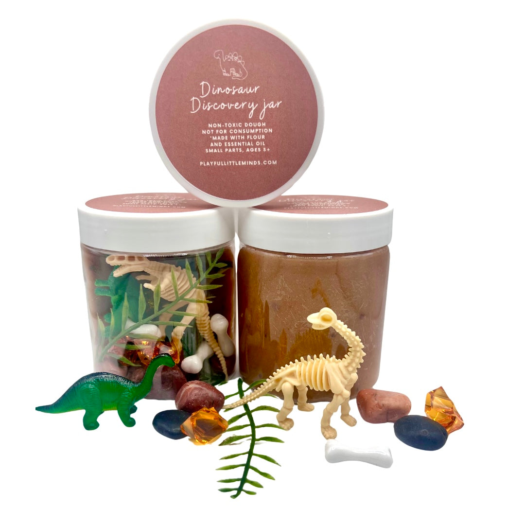 Three jars of Dinosaur Playdough Sensory Kit | Natural Dough with Dino Fossils & Surprises including natural dough, dinosaur skeleton figures, plastic dino toys, and sensory stones arranged with green fern leaves.