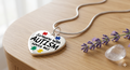 Close-up of a heart-shaped pendant necklace with colorful puzzle piece designs and the text Someone With Autism Loves Me Necklace | Autism Awareness Puzzle Piece Pendant displayed on a wooden surface next to lavender flowers and a clear crystal.