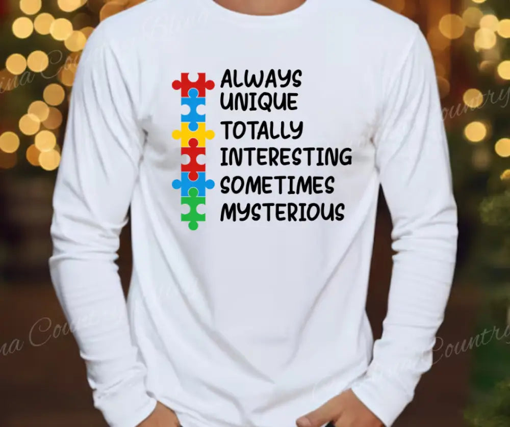 Person wearing a white long-sleeve shirt with colorful puzzle pieces and the words Always Unique Totally Interesting Sometimes Mysterious, showcasing the Lovable AUTISM – Multi Color Tee.