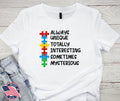 White Lovable AUTISM – Multi Color Tee featuring colorful puzzle pieces and positive words emphasizing uniqueness and mystery, styled casually with denim shorts on a wooden background.