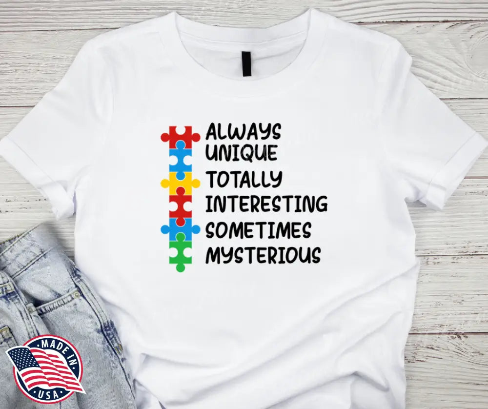 White Lovable AUTISM – Multi Color Tee featuring colorful puzzle pieces and positive words emphasizing uniqueness and mystery, styled casually with denim shorts on a wooden background.