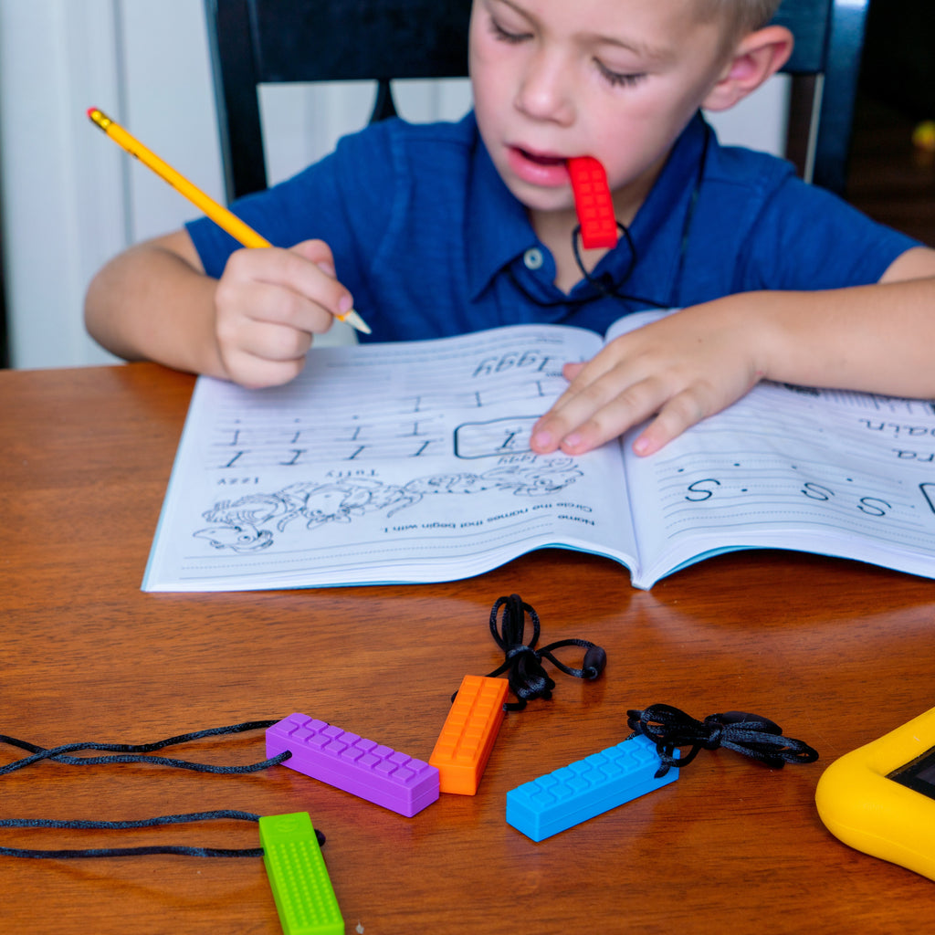 A young boy chewing on a red Chewy Blocks Necklace - Calming Textured Chewy Silicone Pendant for Autism & Oral Motor Special Needs Kids while practicing writing in a workbook at a wooden table with additional colorful chewy pendant necklaces nearby.